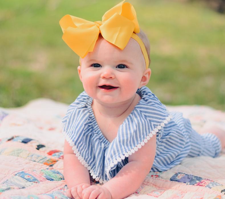 A smiling baby wearing a bow while laying in the grass
